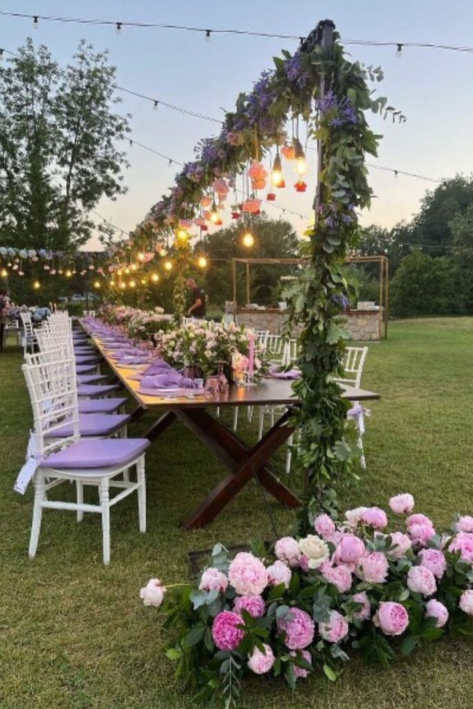 Tavolo imperiale per matrimonio all’aperto con arco floreale, lampadine a luce calda e mise en place nei toni lilla