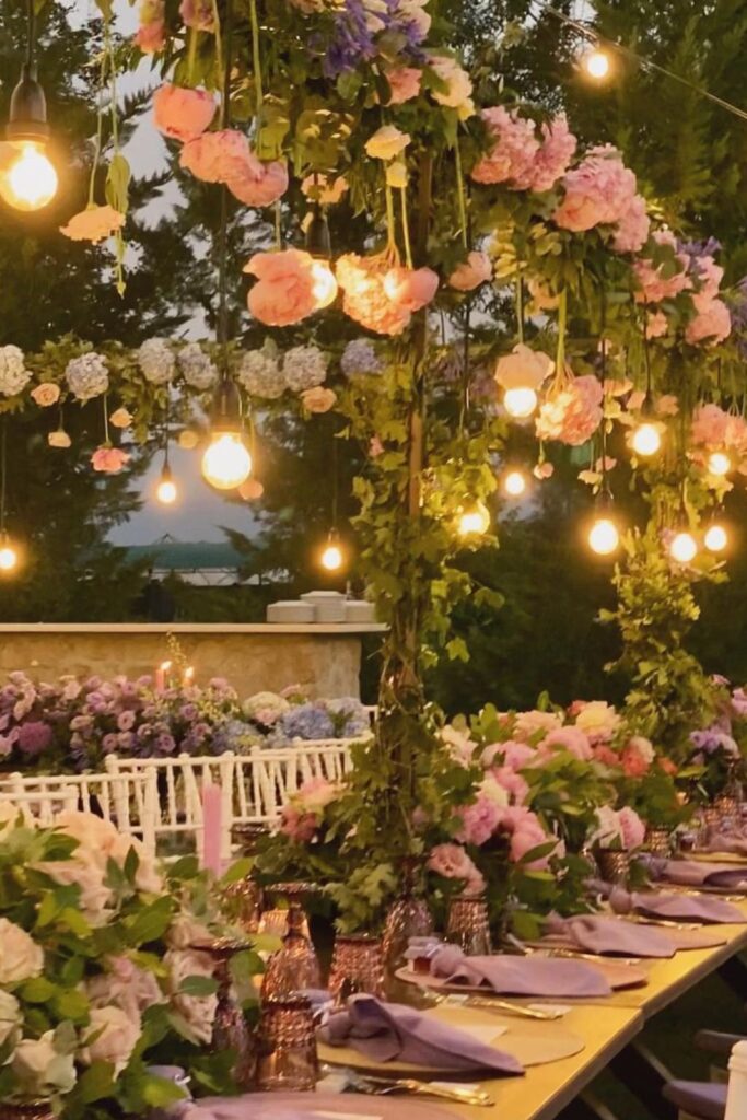 Allestimento floreale sospeso con lampadine a luce calda sopra tavolo imperiale per matrimonio all’aperto