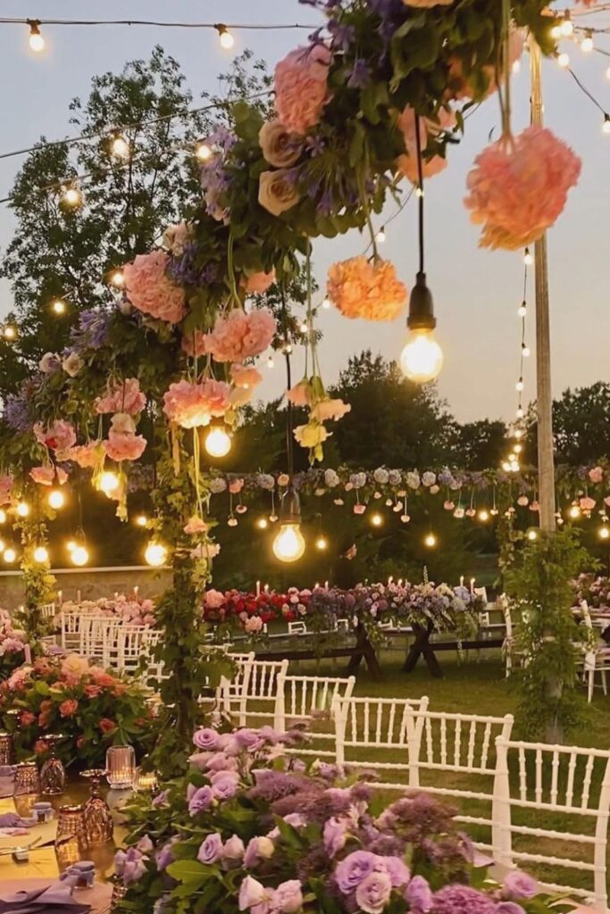 Tavolo imperiale per matrimonio all’aperto con arco floreale, lampadine a luce calda e mise en place nei toni lilla