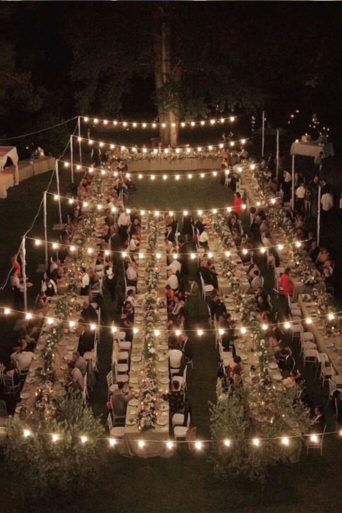 Cena di matrimonio all’aperto vista dall’alto con tavoli imperiali illuminati da luci a filamento sospese
