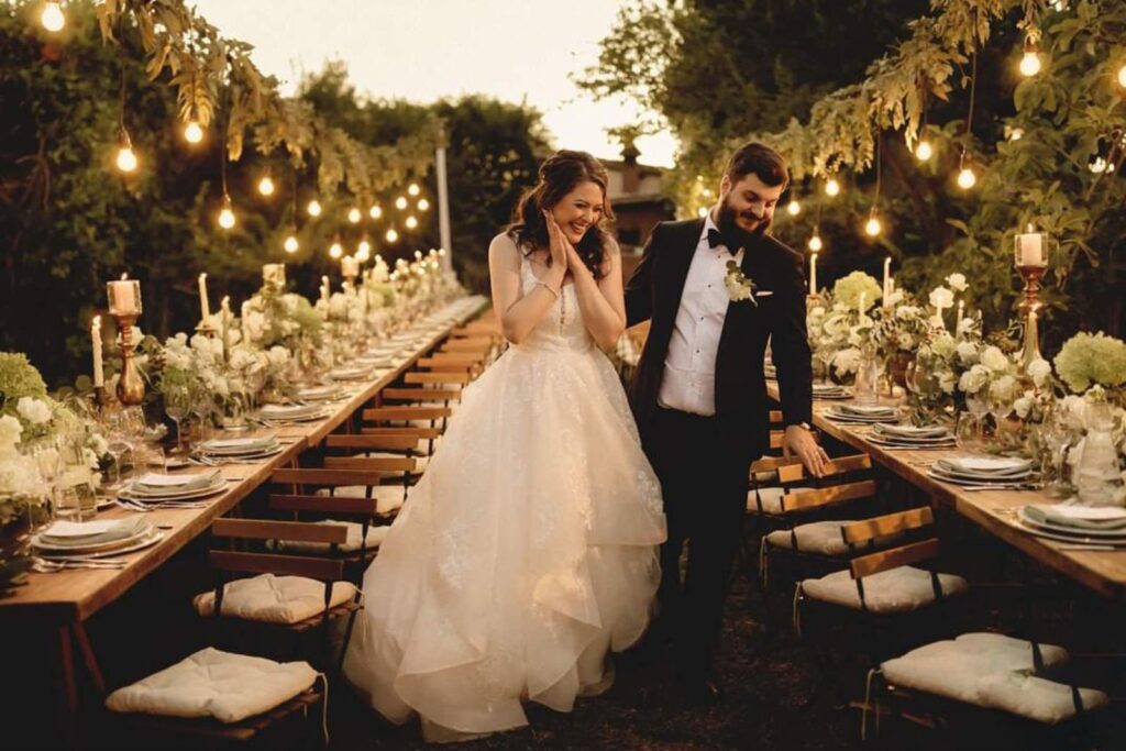 Sposi che camminano tra tavoli imperiali illuminati da lampadine a filamento durante una cena di matrimonio all’aperto
